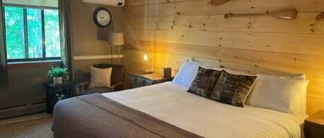 A bedroom with a wooden headboard, a large bed with multiple pillows, a bedside table with a lamp, and a window with curtains.