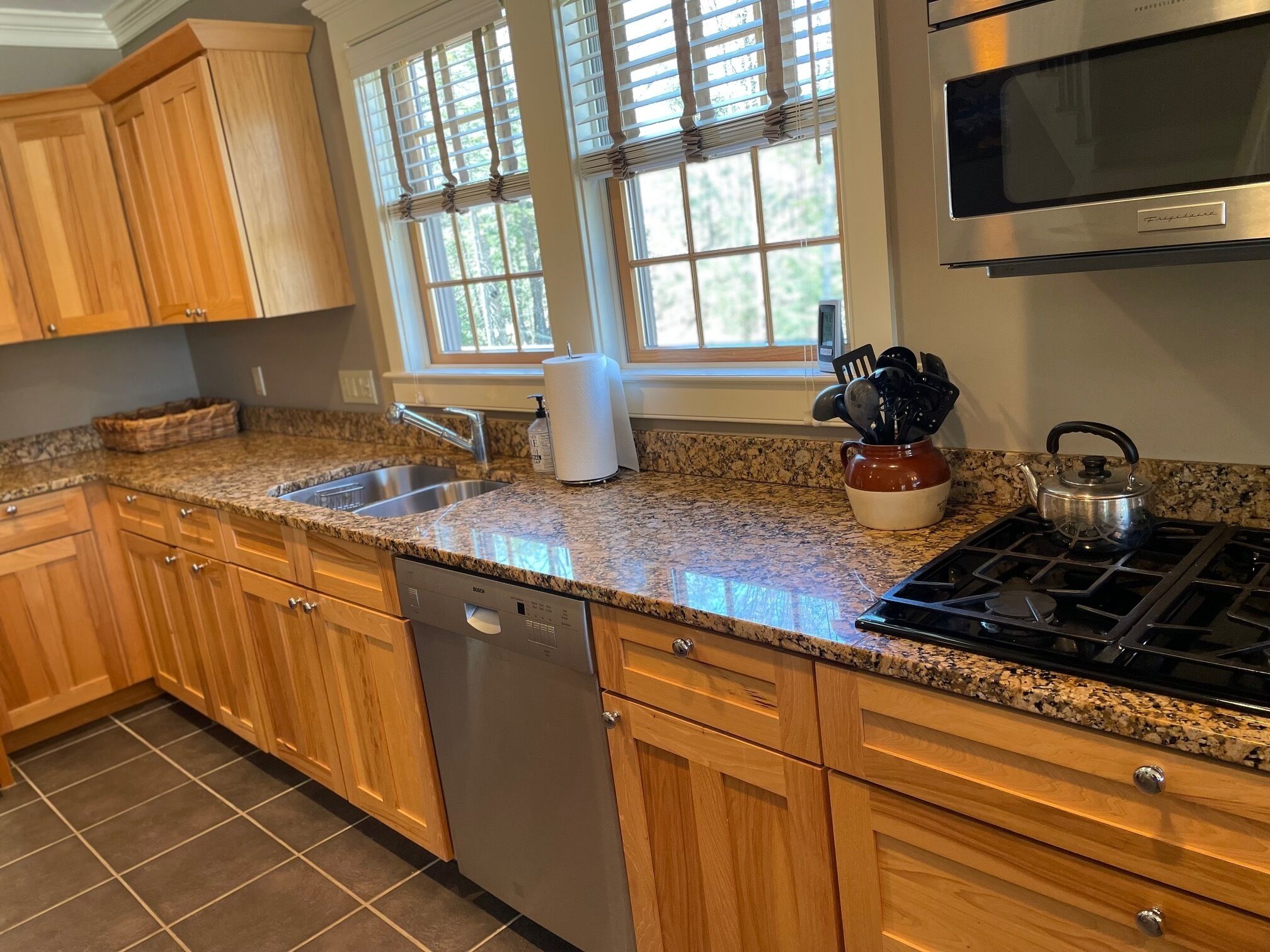 A kitchen with wooden cabinets, a stainless steel dishwasher, a granite countertop, a gas stove, and a window with blinds.