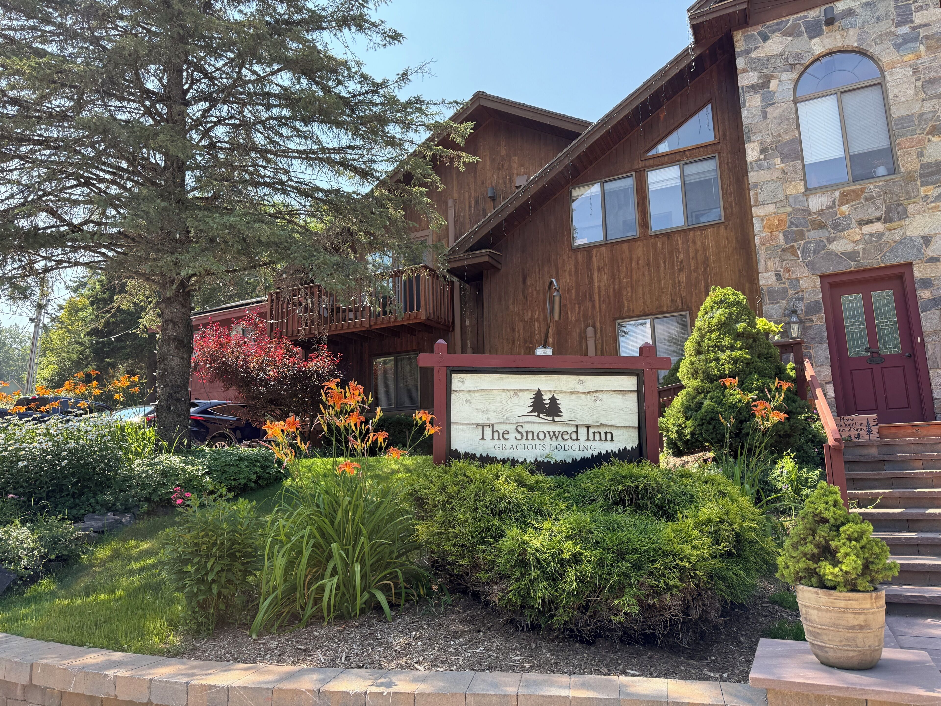 A two-story lodge with a wooden exterior, a signboard for The Snowed Inn, and a well-maintained garden with various plants and flowers.