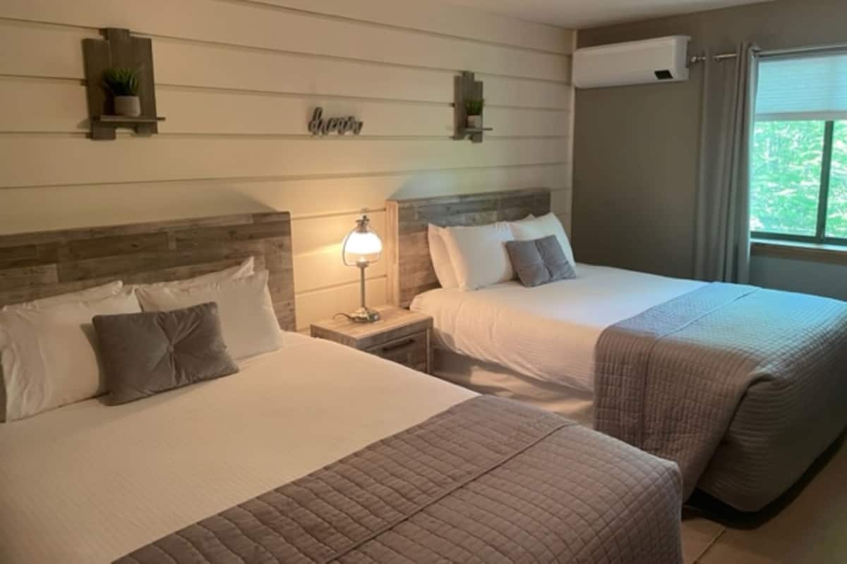 A hotel room with two beds, a nightstand with a lamp, wall-mounted air conditioning, and a window with curtains.