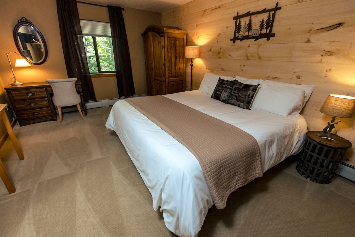 A bedroom with a large bed, a wooden wardrobe, a desk with a chair, and a lamp.