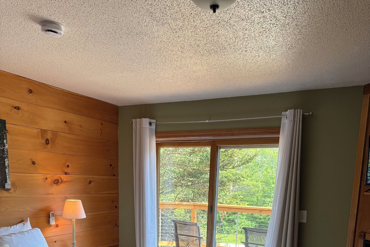 A bedroom with a bed, a bench, a lamp, a ceiling light, and a view of an outdoor deck.