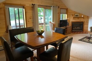 Un interior cálido de cabaña con una mesa de comedor de madera, cuatro sillas, un televisor, una chimenea y ventanas grandes con cortinas.