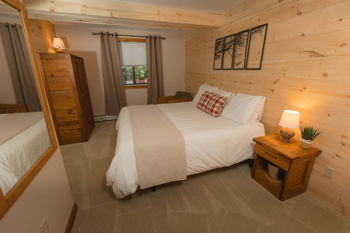 A wooden-paneled bedroom with a bed, a nightstand with a lamp, and a window with curtains.