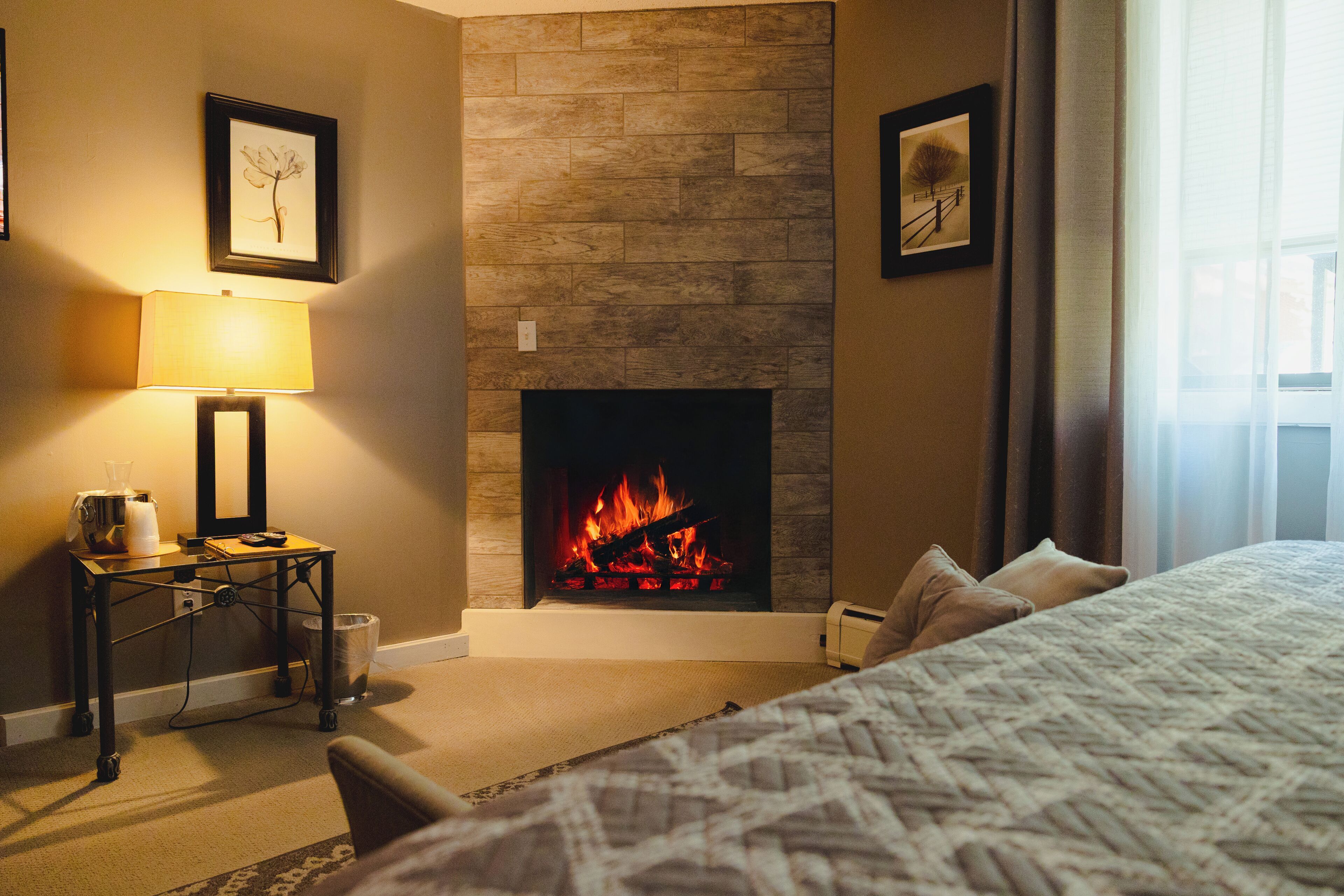 A hotel room with a fireplace, a bed, a bedside table with a lamp, and framed pictures on the wall.