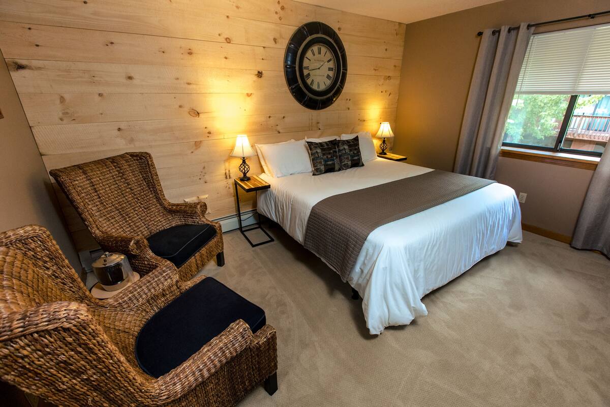 A bedroom with a large bed, two nightstands with lamps, a wicker chair, and a wooden wall with a clock.