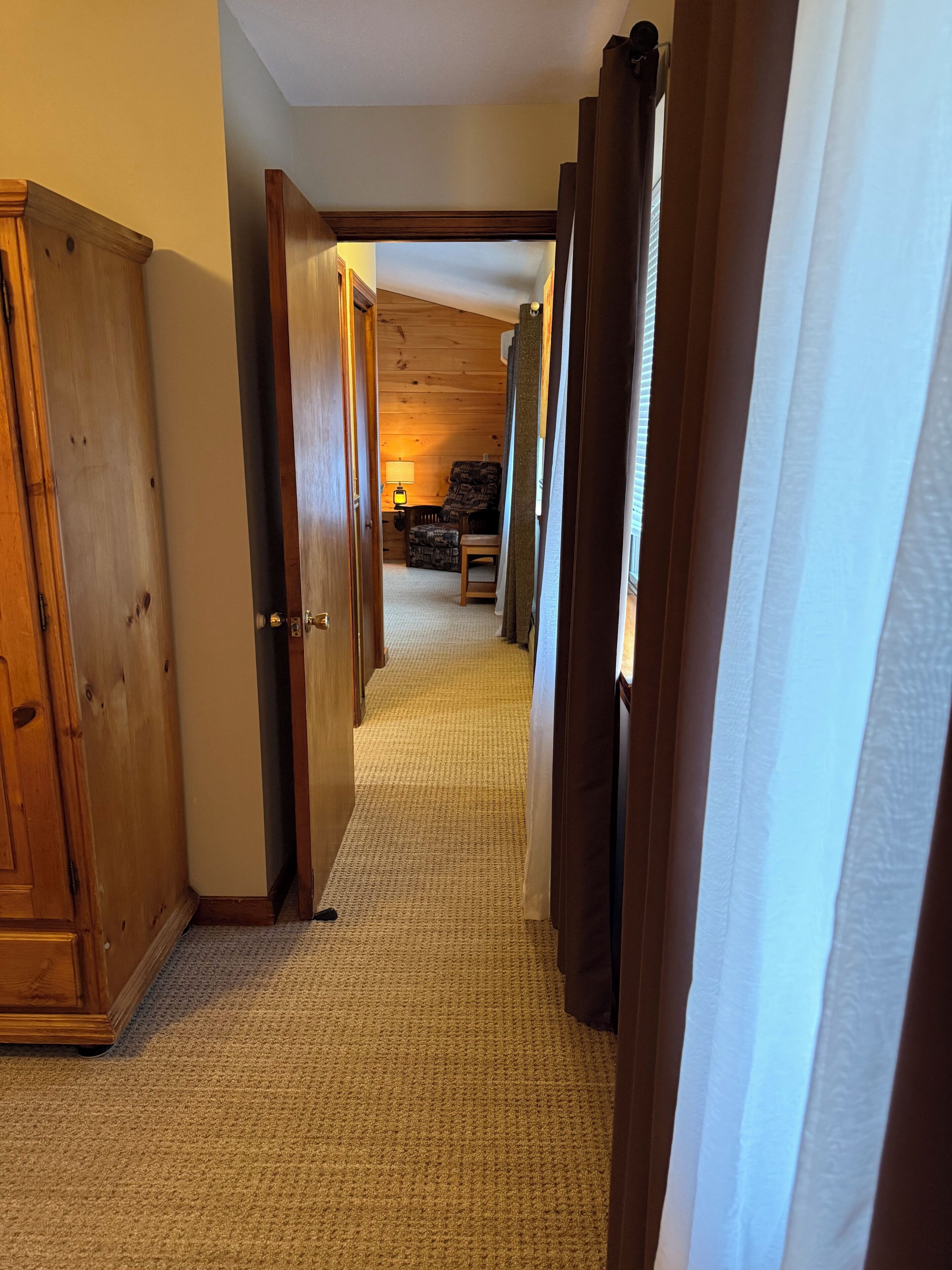 A hallway with wooden doors and a carpeted floor leading to a room with a lamp and a chair.