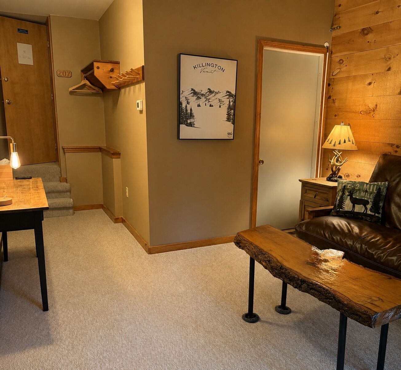 A room with a wooden bench, a brown leather couch, a framed picture on the wall, and a lamp on a wooden nightstand.