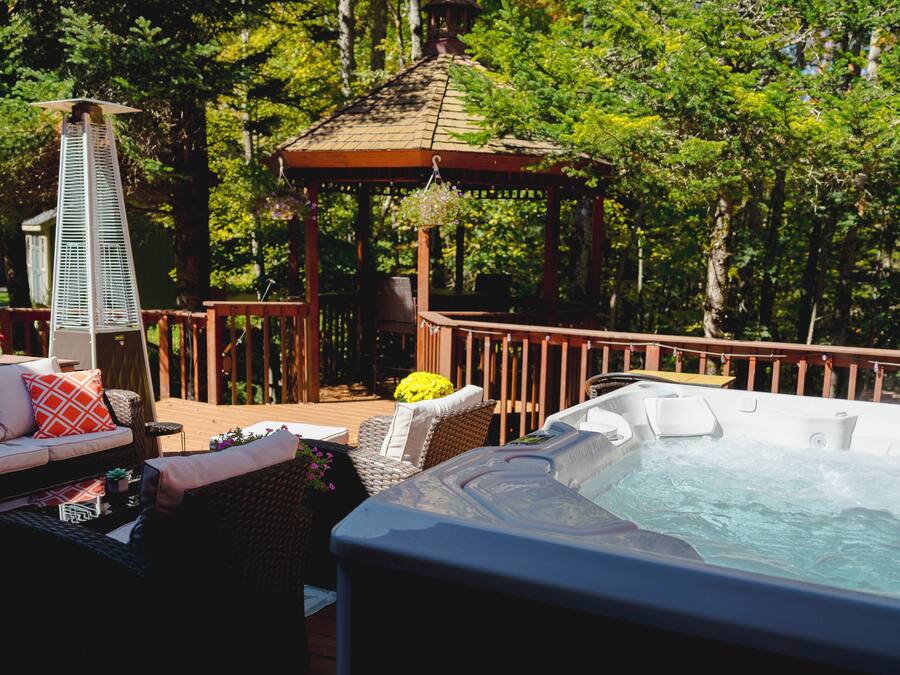 Un bain à remous entouré de meubles de jardin sur une terrasse en bois, avec un belvédère et une végétation luxuriante en arrière-plan.