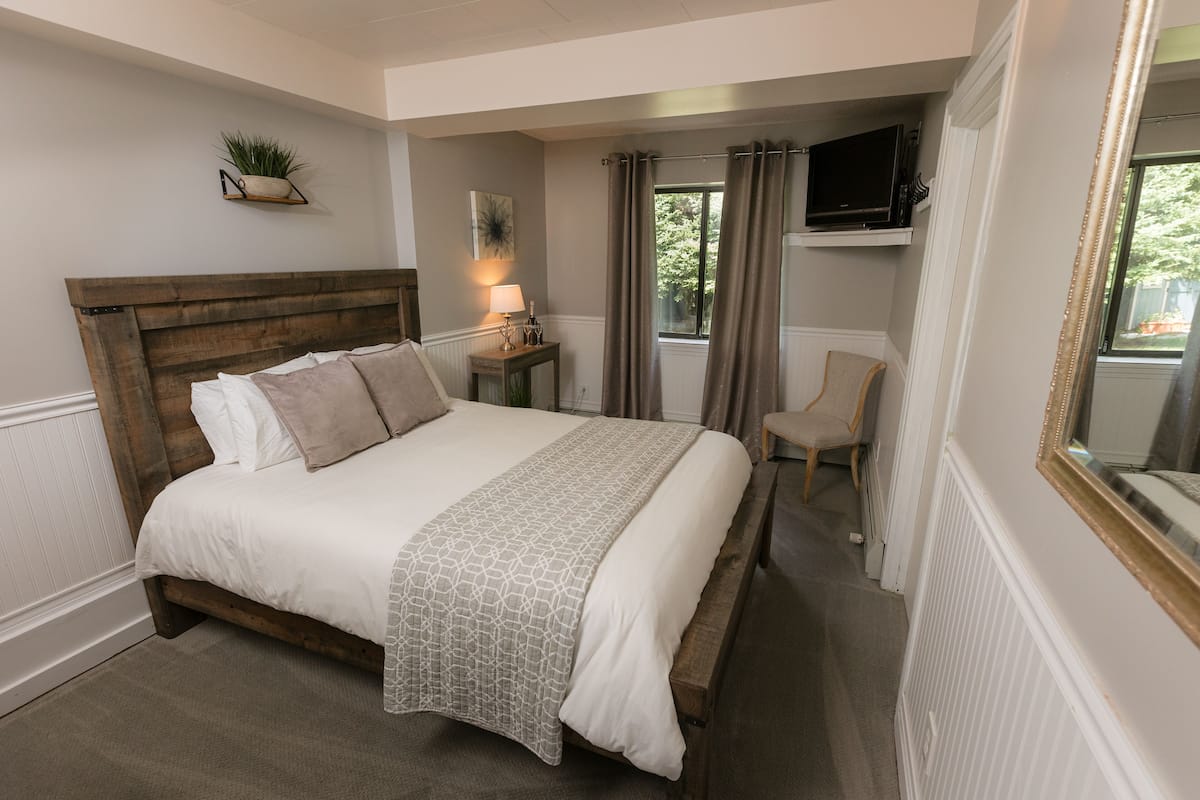 A bedroom with a wooden bed, a bedside table with a lamp, a chair, a TV mounted on the wall, and a window with curtains.