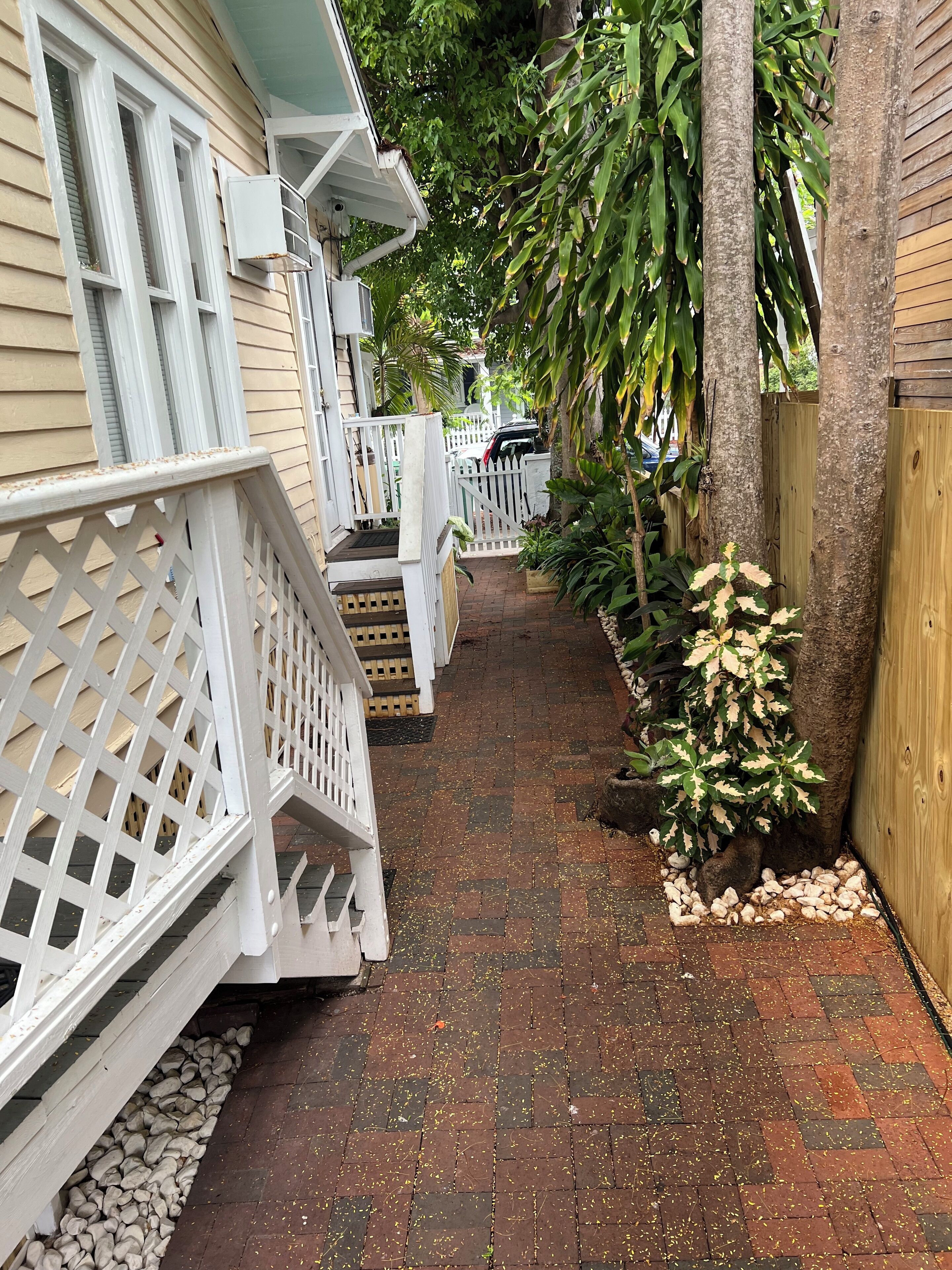 A narrow brick pathway flanked by wooden fences and potted plants, leading to a gated area with a car parked inside.