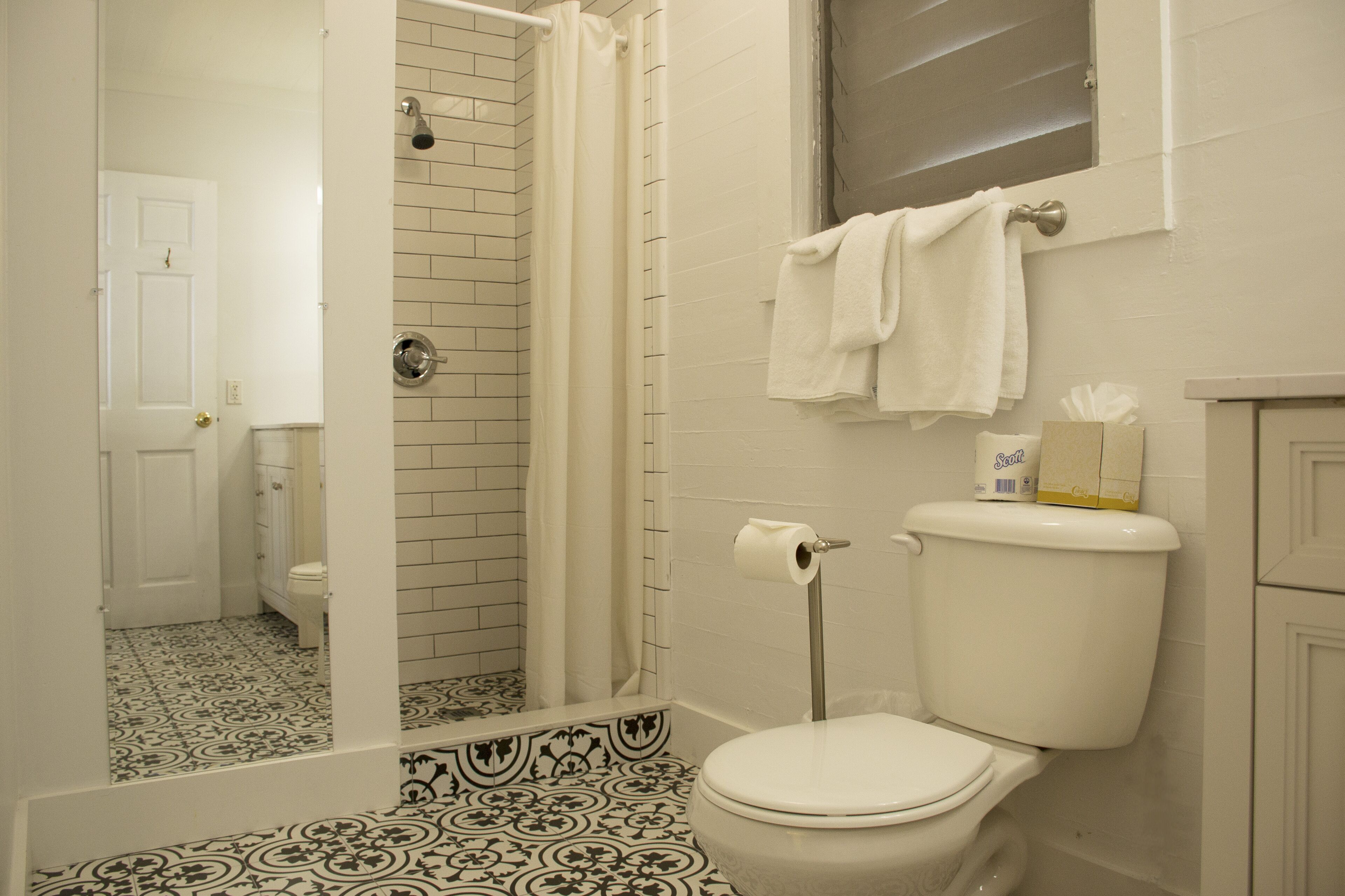 A bathroom with a toilet, a shower with a curtain, a patterned floor, and a towel rack with a box of toilet paper.