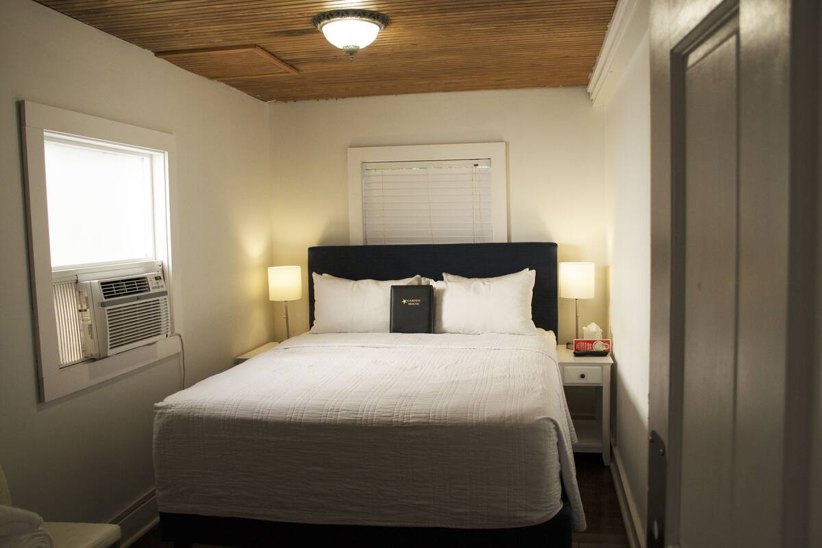 A neatly made bed with white bedding, two bedside lamps, a nightstand with a book and tissues, and a window with blinds.