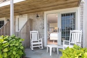 A covered porch with two white rocking chairs and a small table, a wooden staircase, and a door leading to a kitchen area.