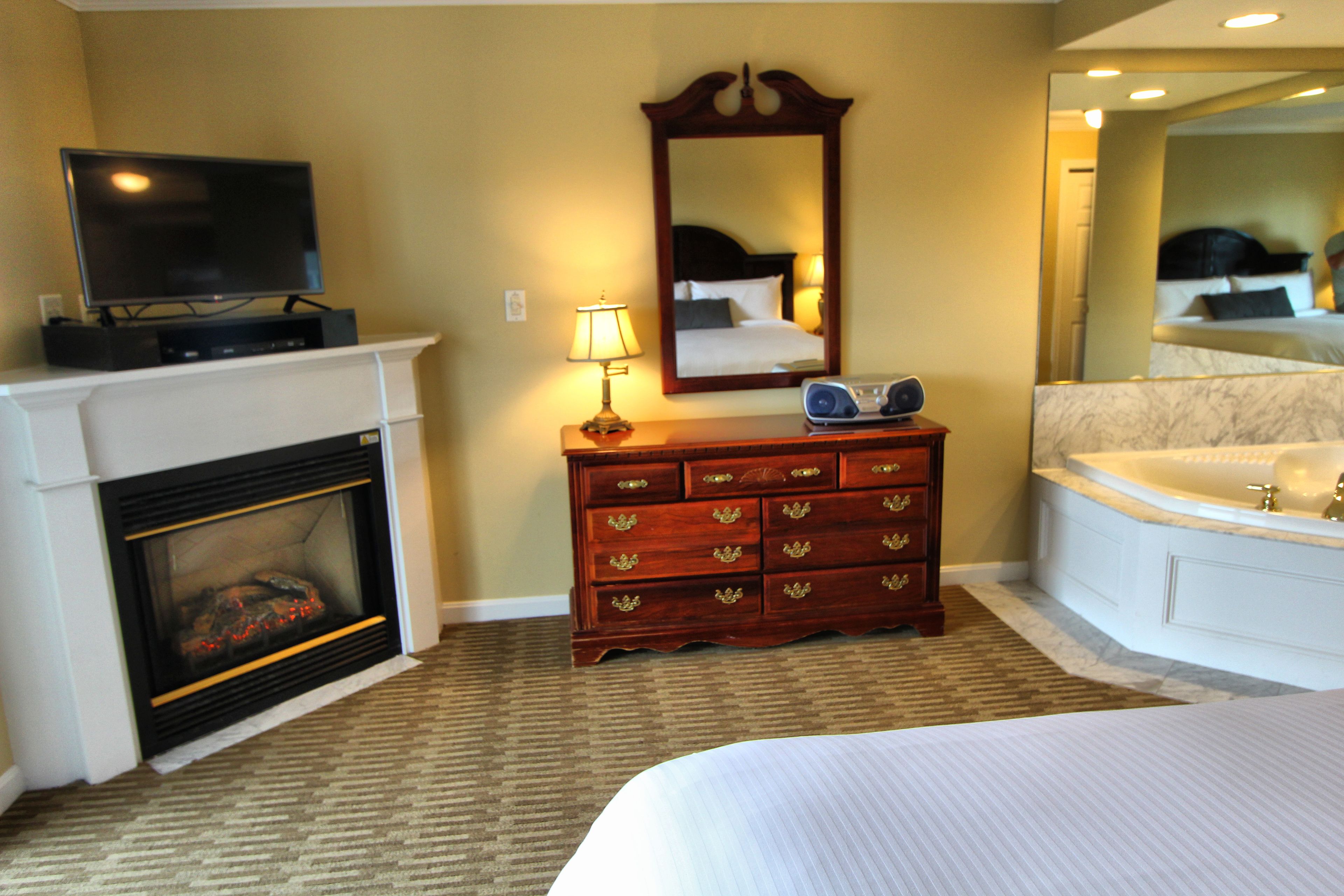 A hotel room with a fireplace, a wooden dresser, a television, a lamp, a mirror, and a large bathtub.