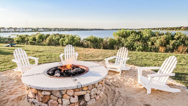 A stone fire pit with four Adirondack chairs around it on a sandy beach with a view of a lake and distant houses. - InnSeason Resorts Surfside (East Falmouth)