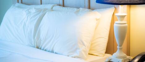 Una cama bien tendida con sábanas blancas, un cabecero de madera y una mesita de noche con una lámpara y un teléfono.