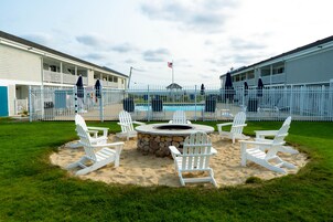 A pool area with white Adirondack chairs and a stone fire pit surrounded by sand.