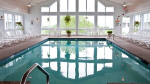 An indoor swimming pool with clear views through large windows, white lounge chairs, and potted plants. - InnSeason Resorts Surfside (East Falmouth)