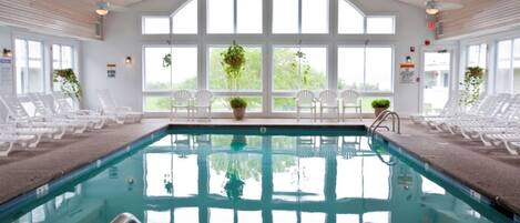 Une piscine intérieure offrant une vue dégagée grâce à de grandes fenêtres, des fauteuils de relaxation blancs et des plantes en pot.