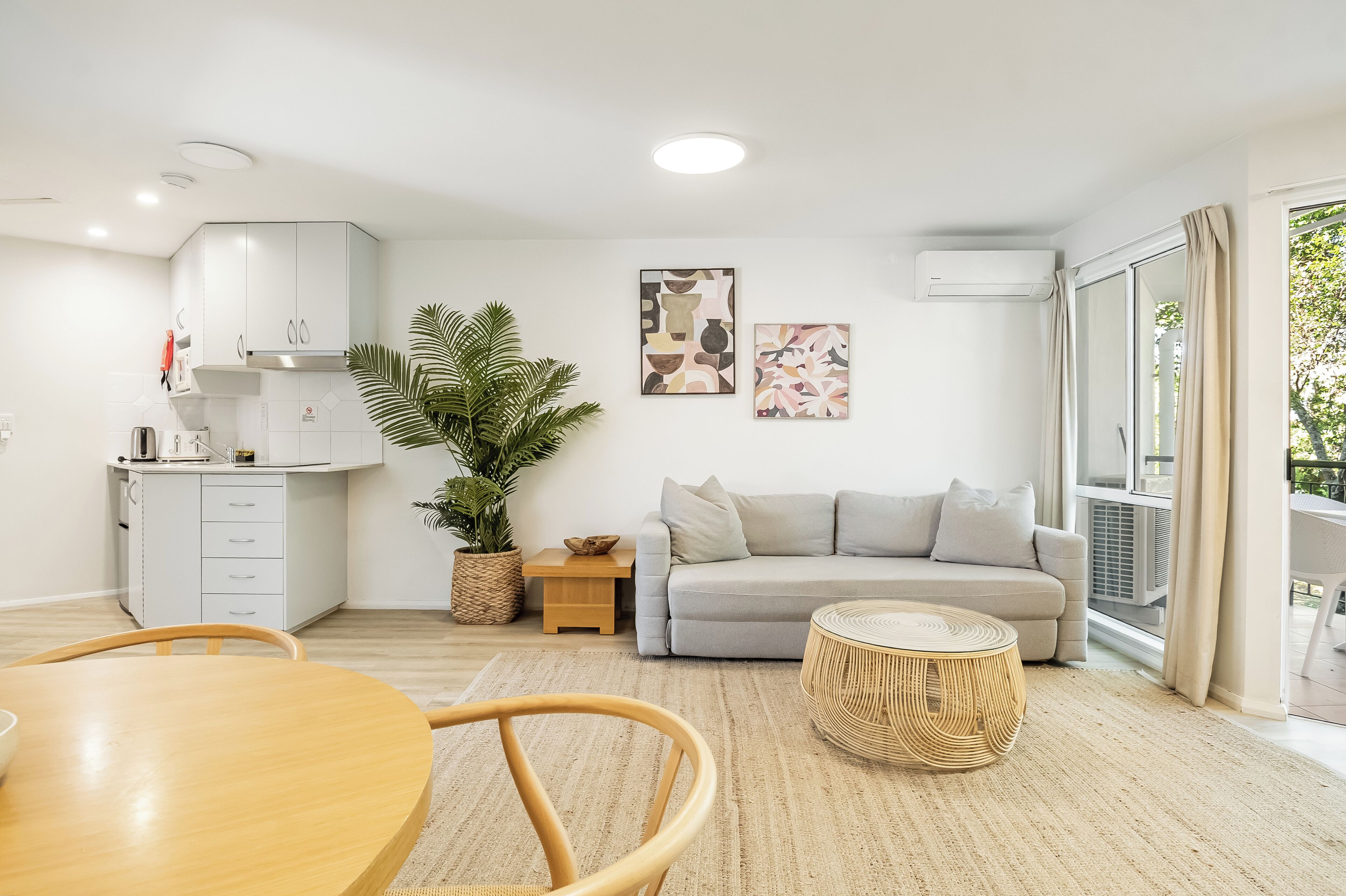 Una sala moderna con un sofá gris, una mesa redonda de madera y una mesa de comedor con sillas. Se observa una zona de cocina con armarios blancos y una planta en maceta.