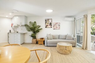 Una sala moderna con un sofá gris, una mesa redonda de madera y una mesa de comedor con sillas. Se observa una zona de cocina con armarios blancos y una planta en maceta.