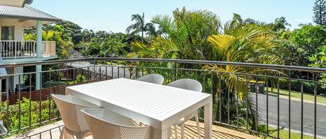 A balcony with a white table and chairs, surrounded by greenery and a view of a road.