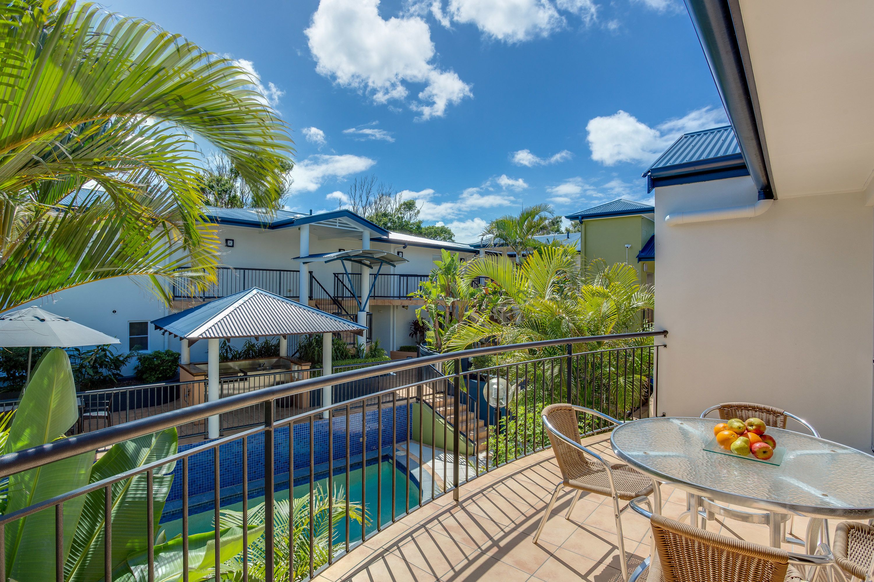 A balcony with a glass table and chairs, a pool, and a view of neighboring buildings.