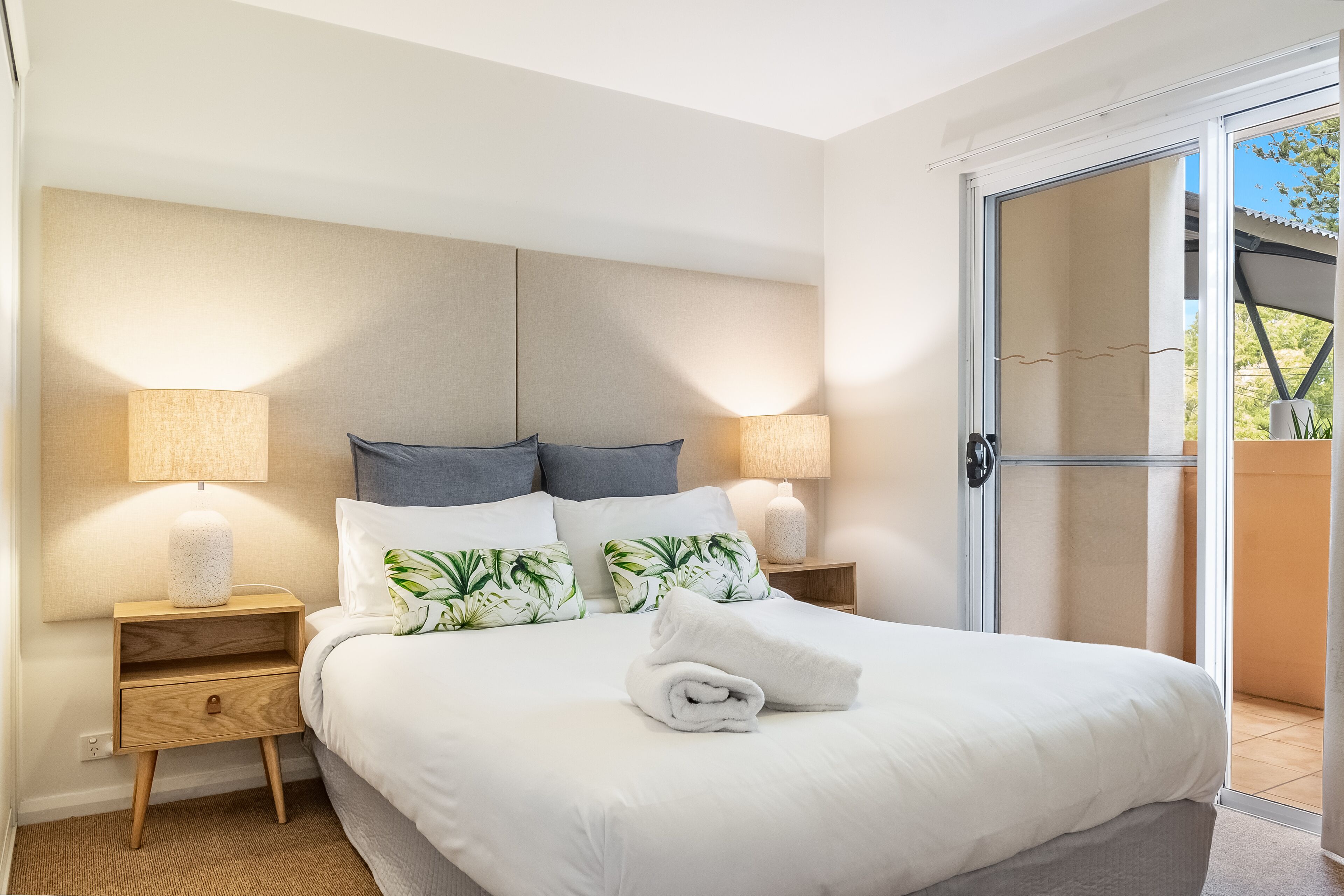 A neatly made bed with white linens and a rolled towel, flanked by two bedside tables with lamps, a wooden nightstand, and a large sliding glass door leading to an outdoor area.