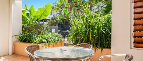 A patio with a round glass table and wicker chairs, surrounded by potted plants and a view of buildings.