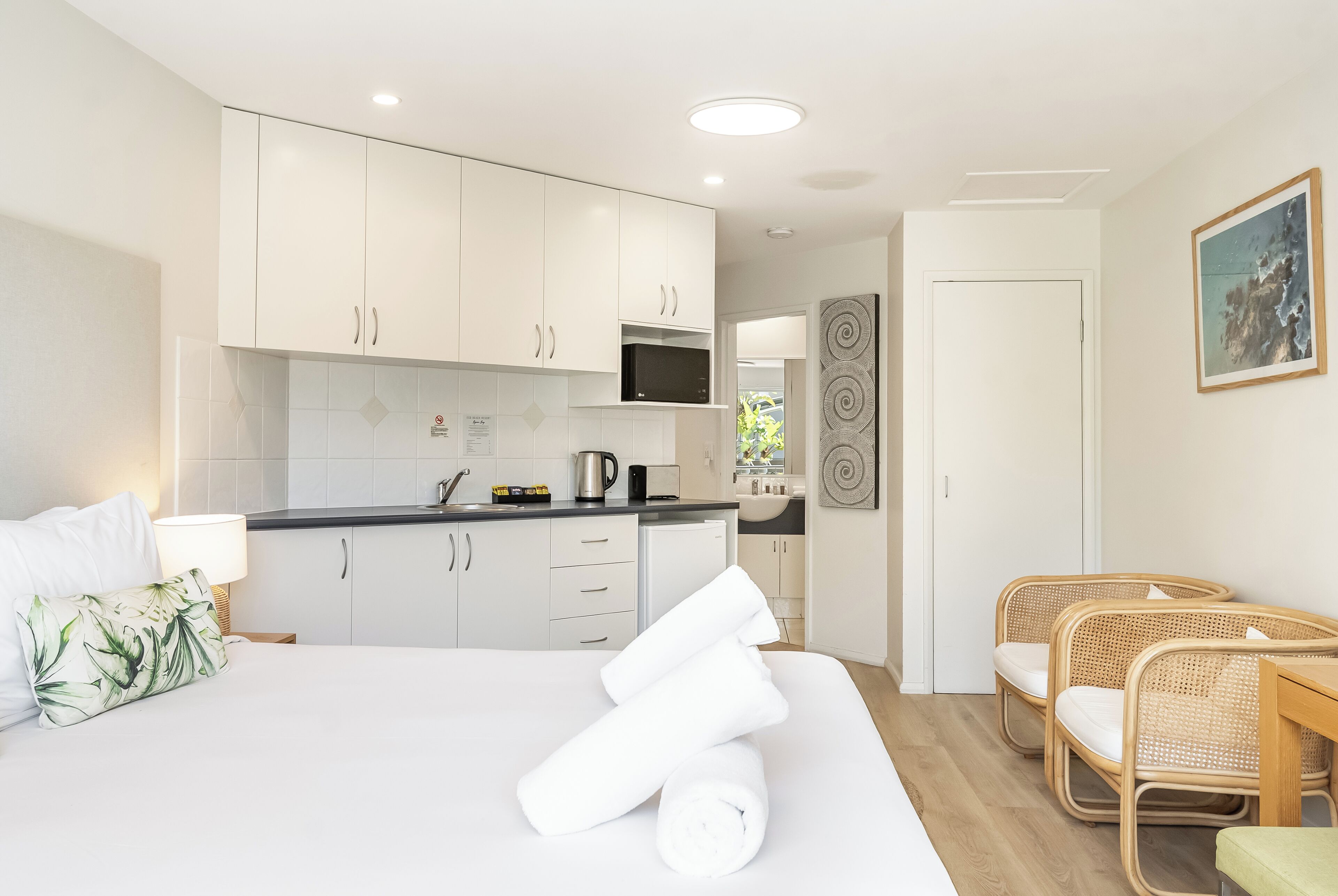 Une chambre d’hôtel moderne comprenant un lit, un coin repas avec des chaises en osier, une kitchenette et une salle de bain visible à travers une porte ouverte.