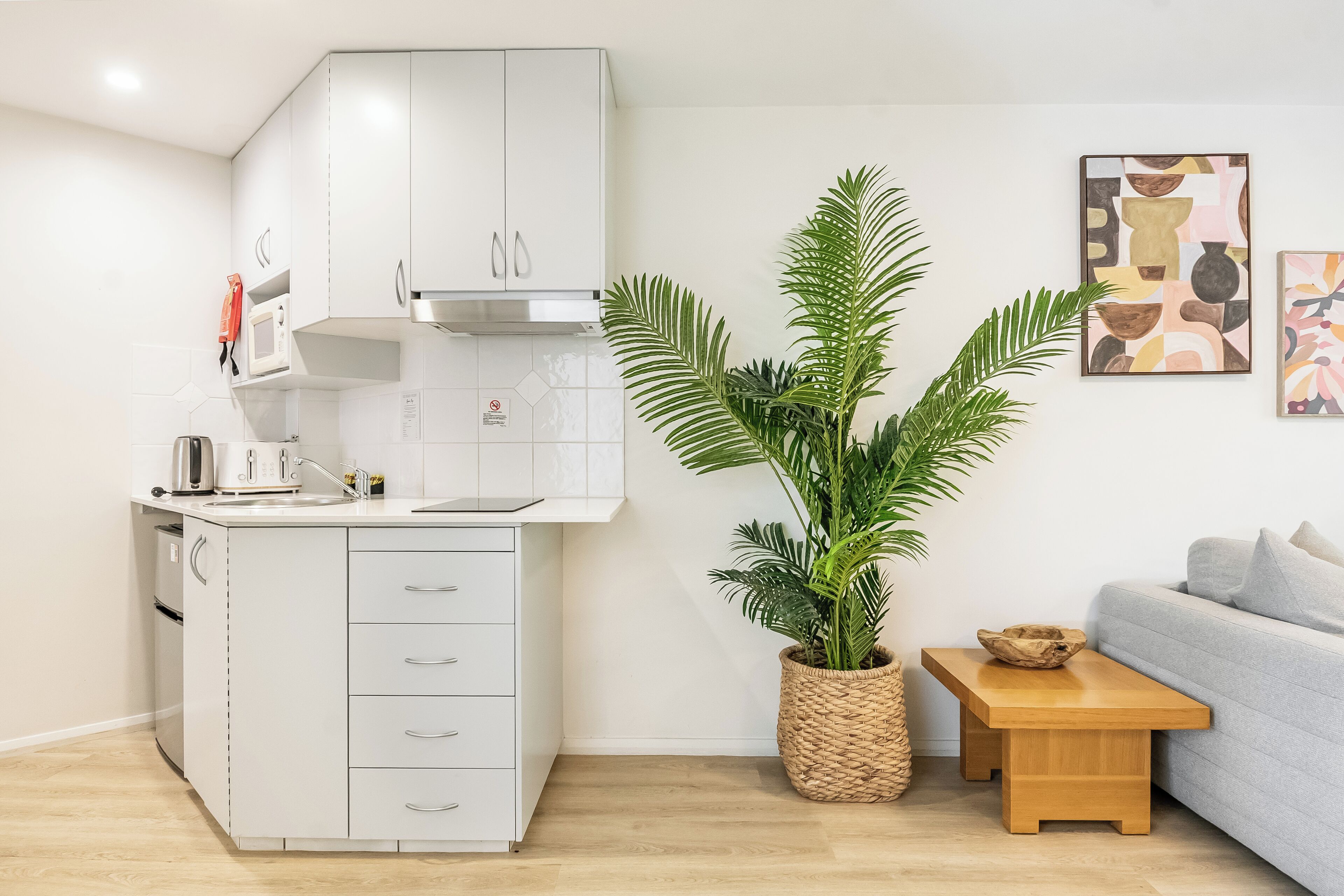 Una cocina moderna con armarios blancos, un microondas y un lavamanos. Hay una mesa de madera con un cuenco y una planta en maceta. Un sofá gris está apoyado contra la pared.