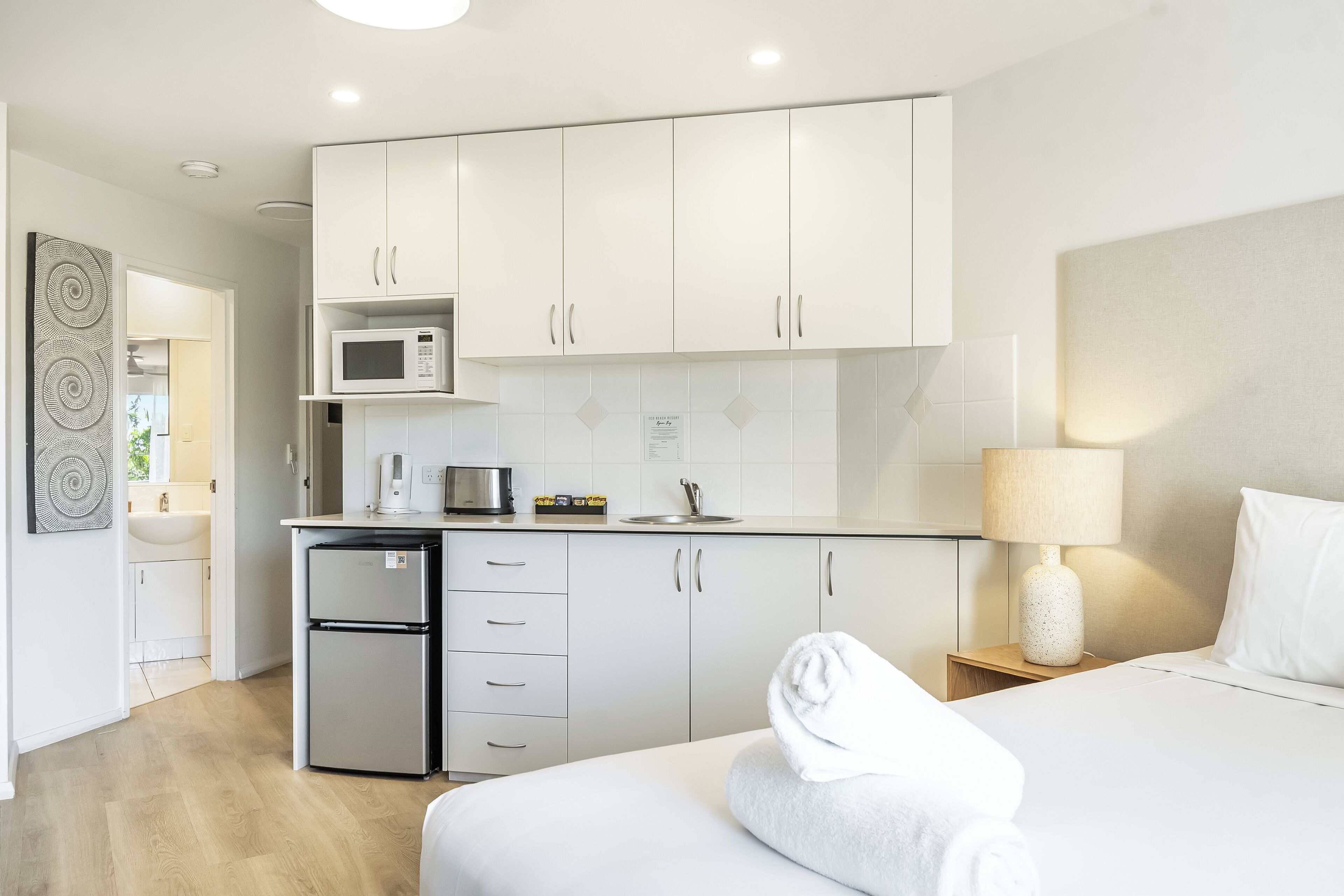 Una habitación de hotel moderna con una cama, una mesita de noche con lámpara, una kitchenette con microondas y un refrigerador.