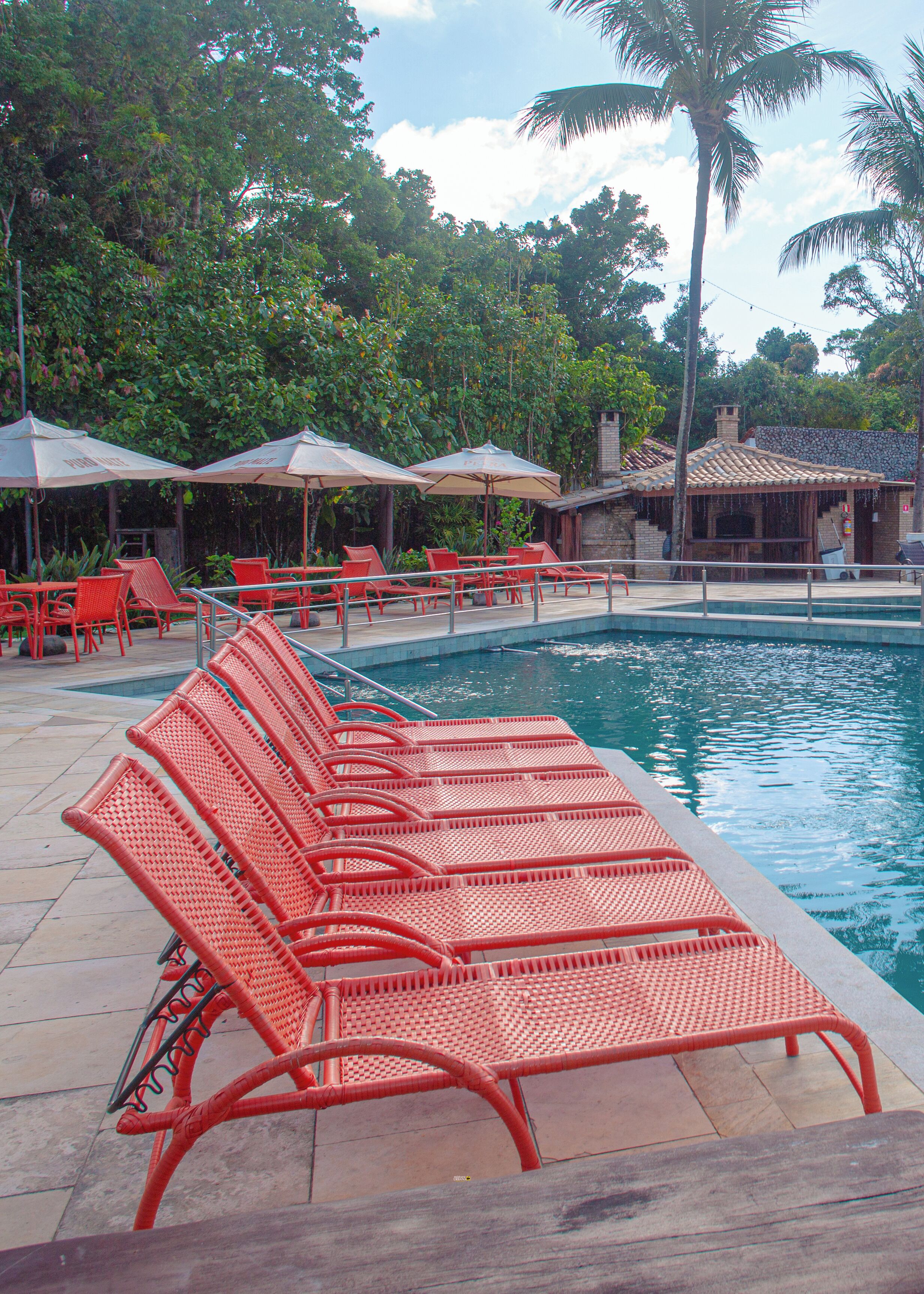 A poolside area with red lounge chairs and umbrellas, surrounded by lush greenery and a stone building.