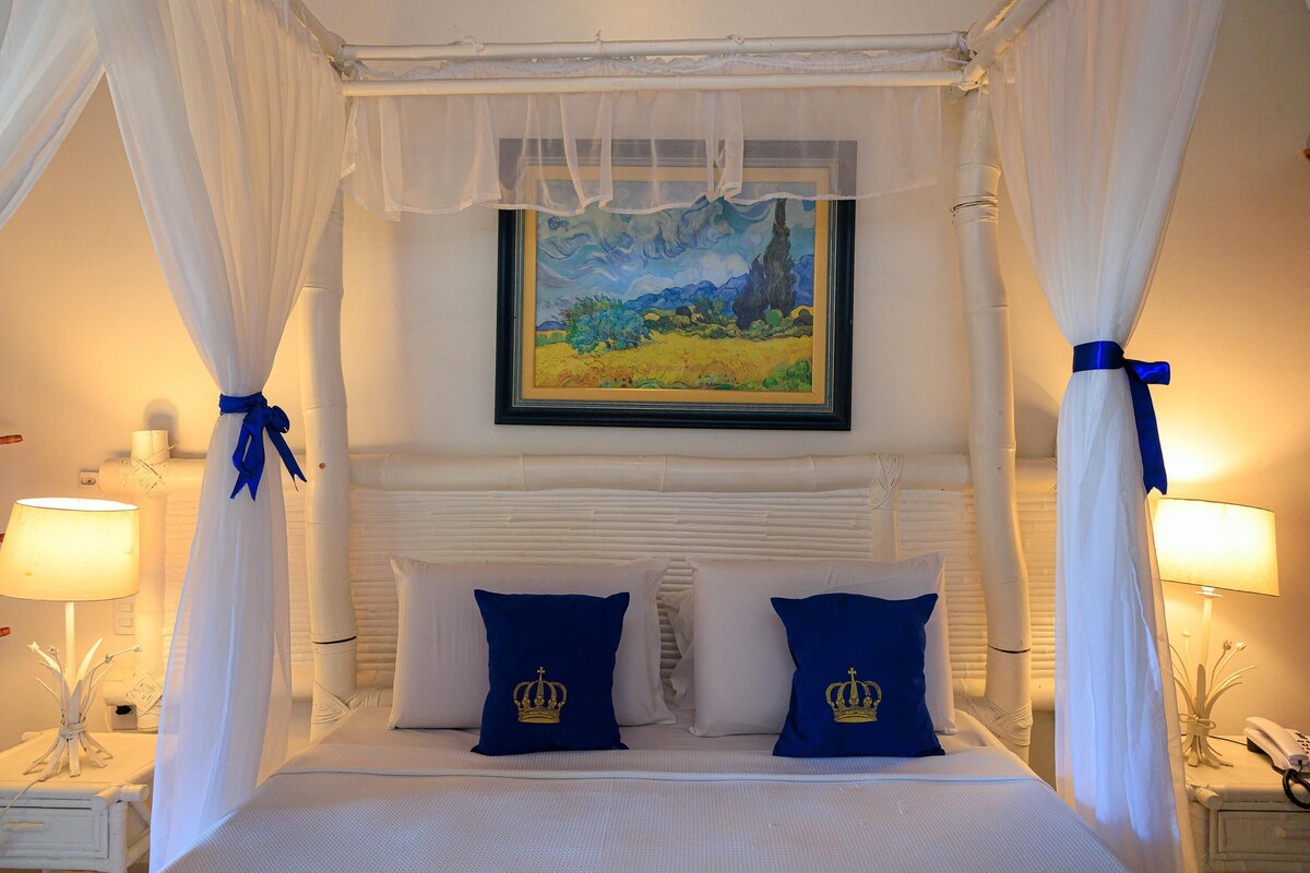 Una cama con ropa blanca, un cuadro enmarcado encima y un dosel decorado con cintas azules.