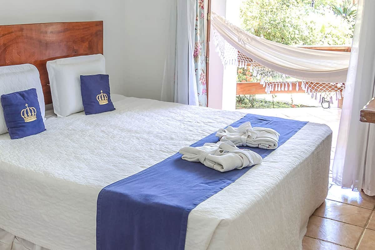 A hotel room with a wooden headboard, a bed with white bedding and blue accents, a wooden chair, and a view of greenery outside.
