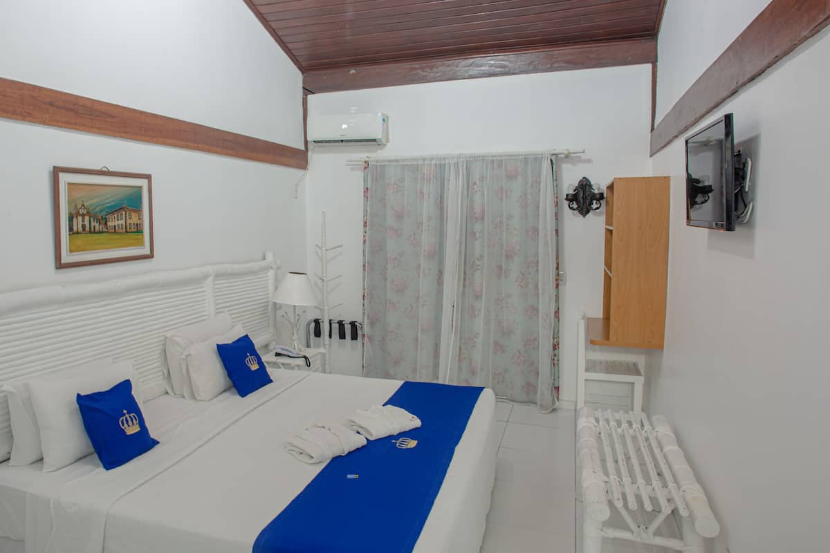 A hotel room with a bed, a white radiator, a painting on the wall, and an air conditioning unit.