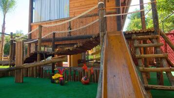 Una estructura de juego de madera con un puente de cuerda y un tobogán, rodeada por una cerca y follaje.