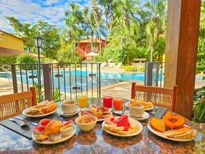 Mesa de café da manhã com diversos pratos, piscina e palmeiras ao fundo.