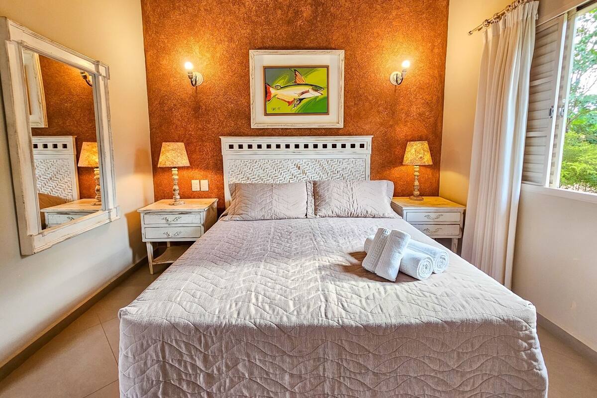 A bedroom with a large bed, two bedside tables, a headboard with a woven pattern, a wall-mounted painting of a fish, and a window with curtains.