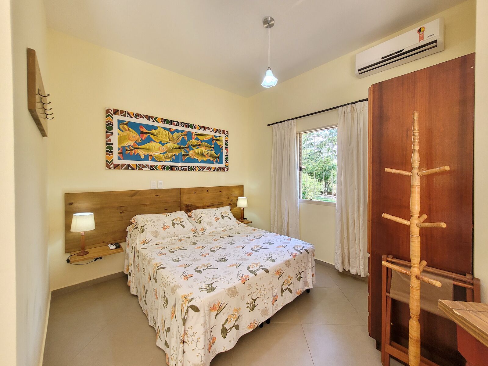 A bedroom with a wooden bed, a wall-mounted air conditioner, a window with curtains, and a decorative painting.