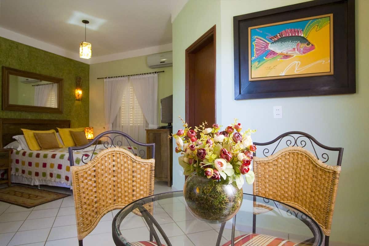 A room with a bed, a dining table with chairs, a vase with flowers, and a framed fish painting on the wall.