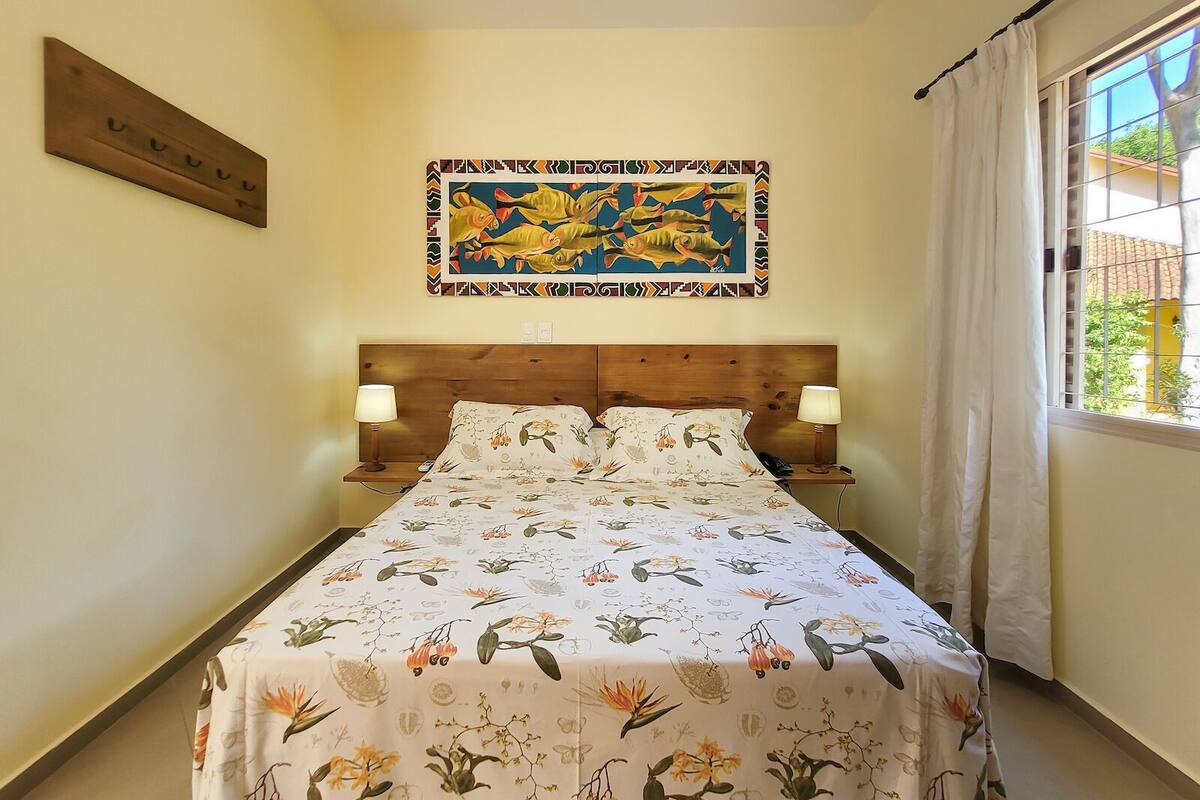 A bedroom with a bed, two bedside tables, a nightstand, a wall-mounted artwork, and a window with curtains.