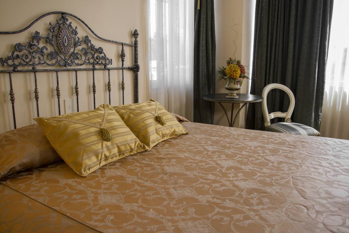 A neatly made bed with a patterned bedspread and two decorative pillows, a metal headboard, a small round table with a vase of flowers, and a chair with a striped cushion.