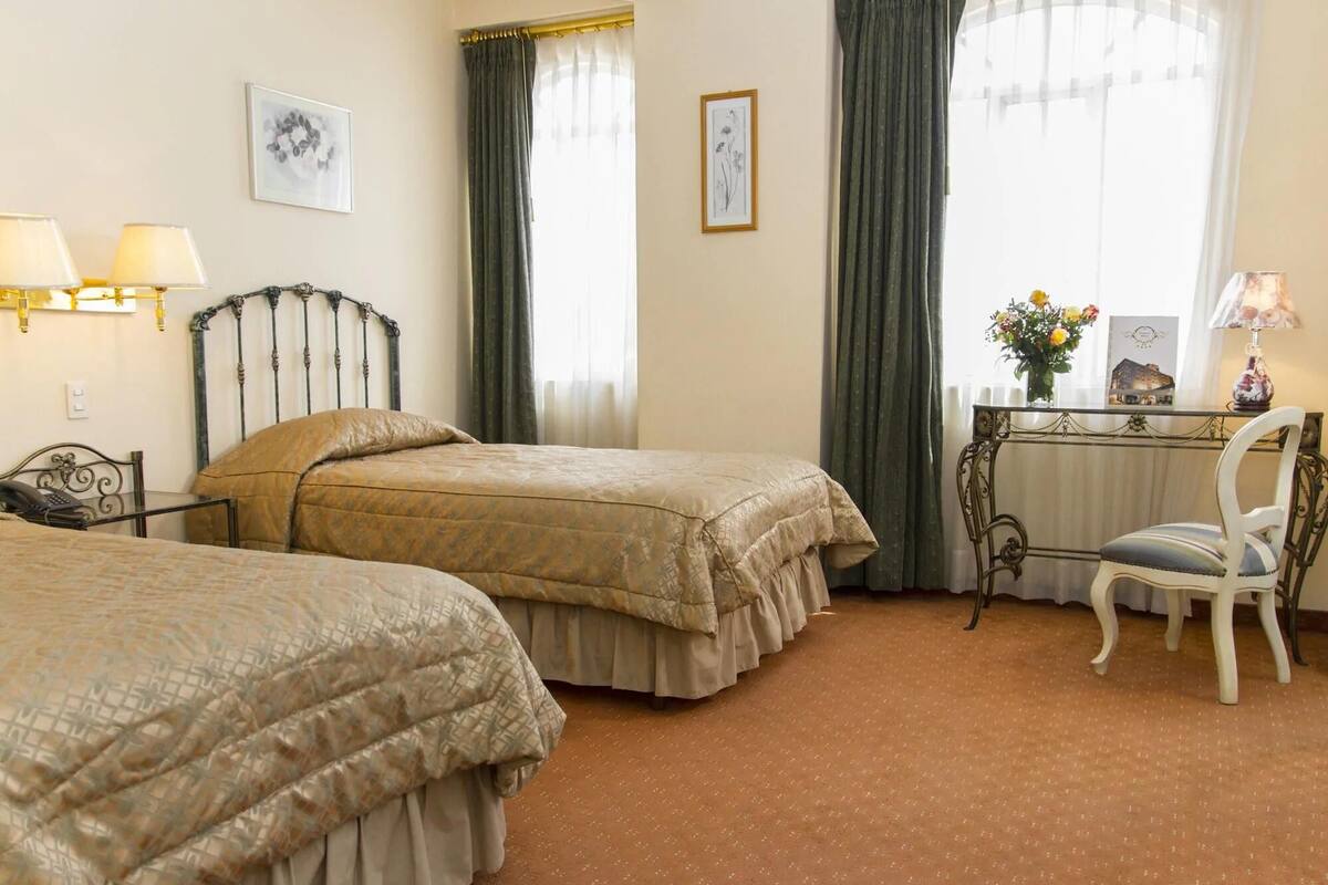 A hotel room with two beds, a desk with a chair, a lamp, and a window with curtains.