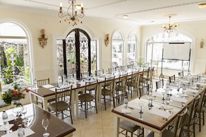 Uma sala de jantar formal com mesas compridas preparadas para uma refeição, com lustres e grandes janelas.