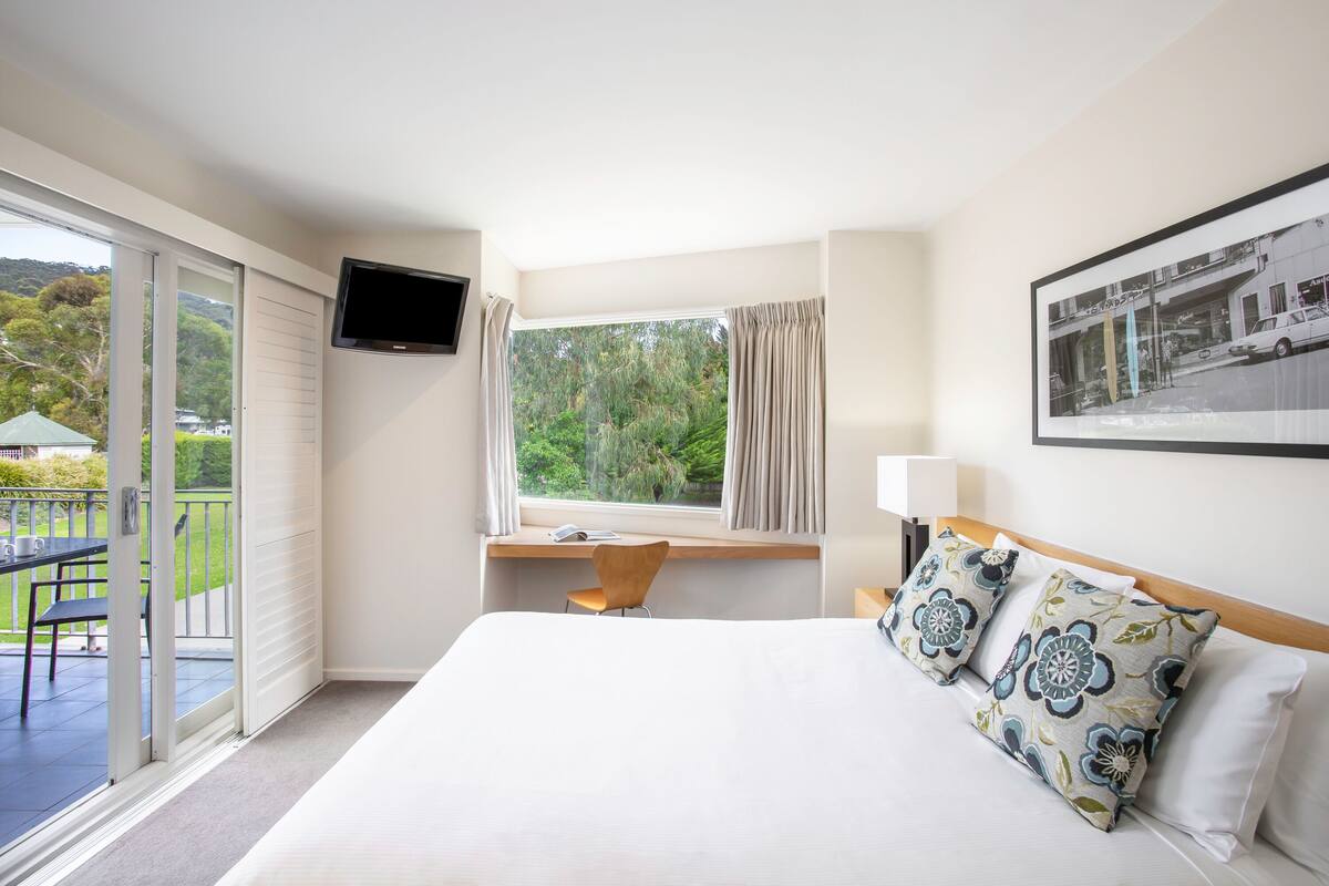 Habitación de hotel con cama, escritorio con silla, televisor montado en la pared, ventana con cortinas y balcón con mesa y sillas.