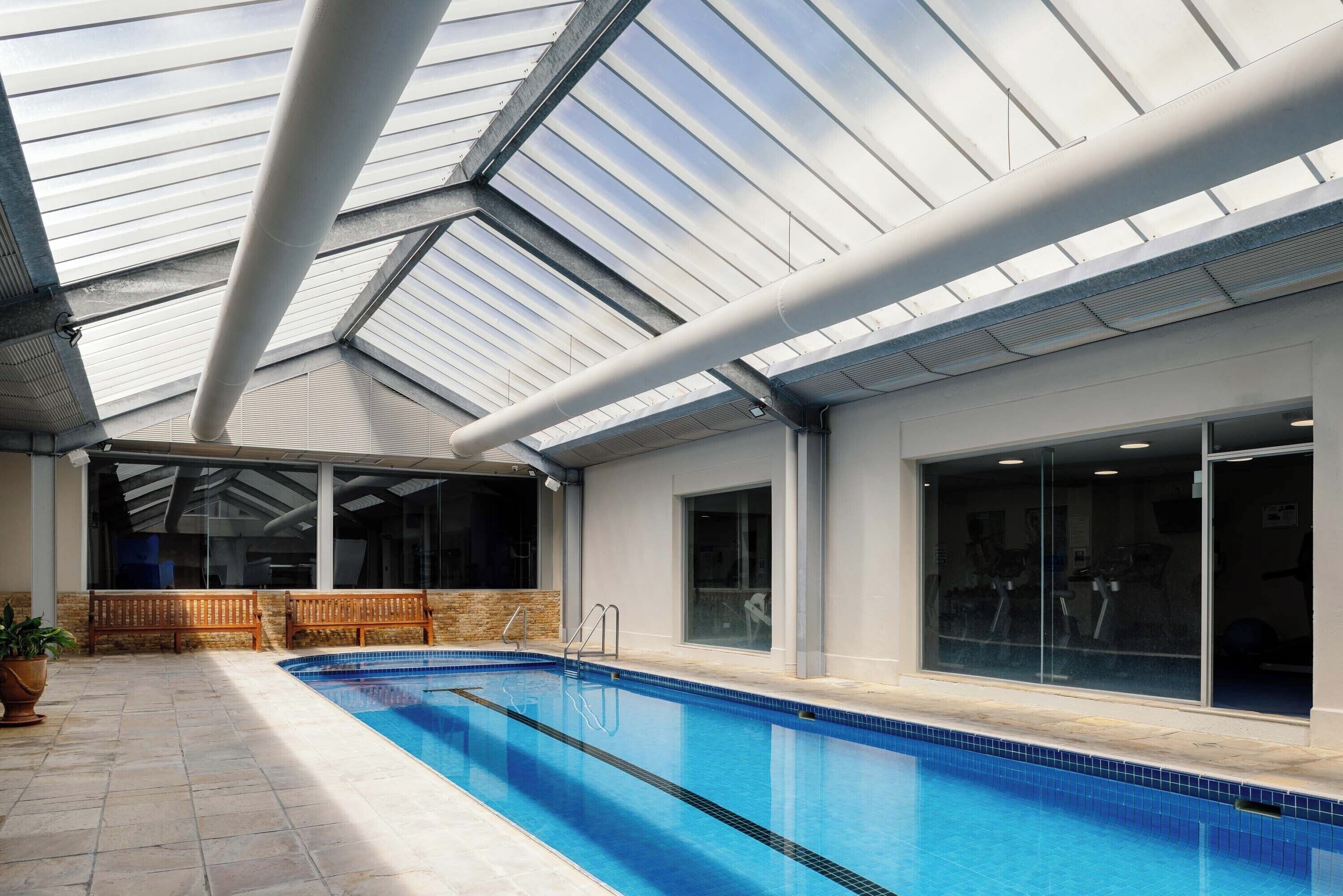 An indoor swimming pool with a skylight roof, wooden benches, and exercise equipment in the background.