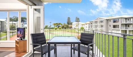 A balcony with a table and chairs, a television on a stand, and a view of a sports field and buildings in the background.