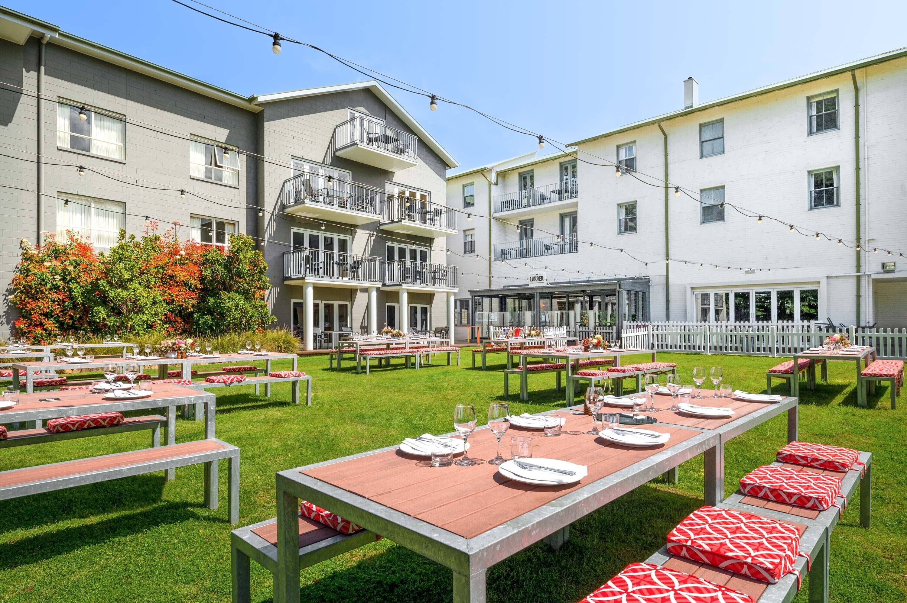 Un área de comedor al aire libre con mesas y bancos, rodeada de edificios con balcones y una cerca blanca de piquetes.